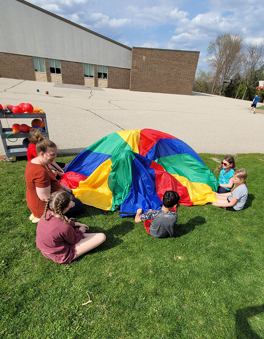 Kids sitting outside playing with a rainbow parachute.